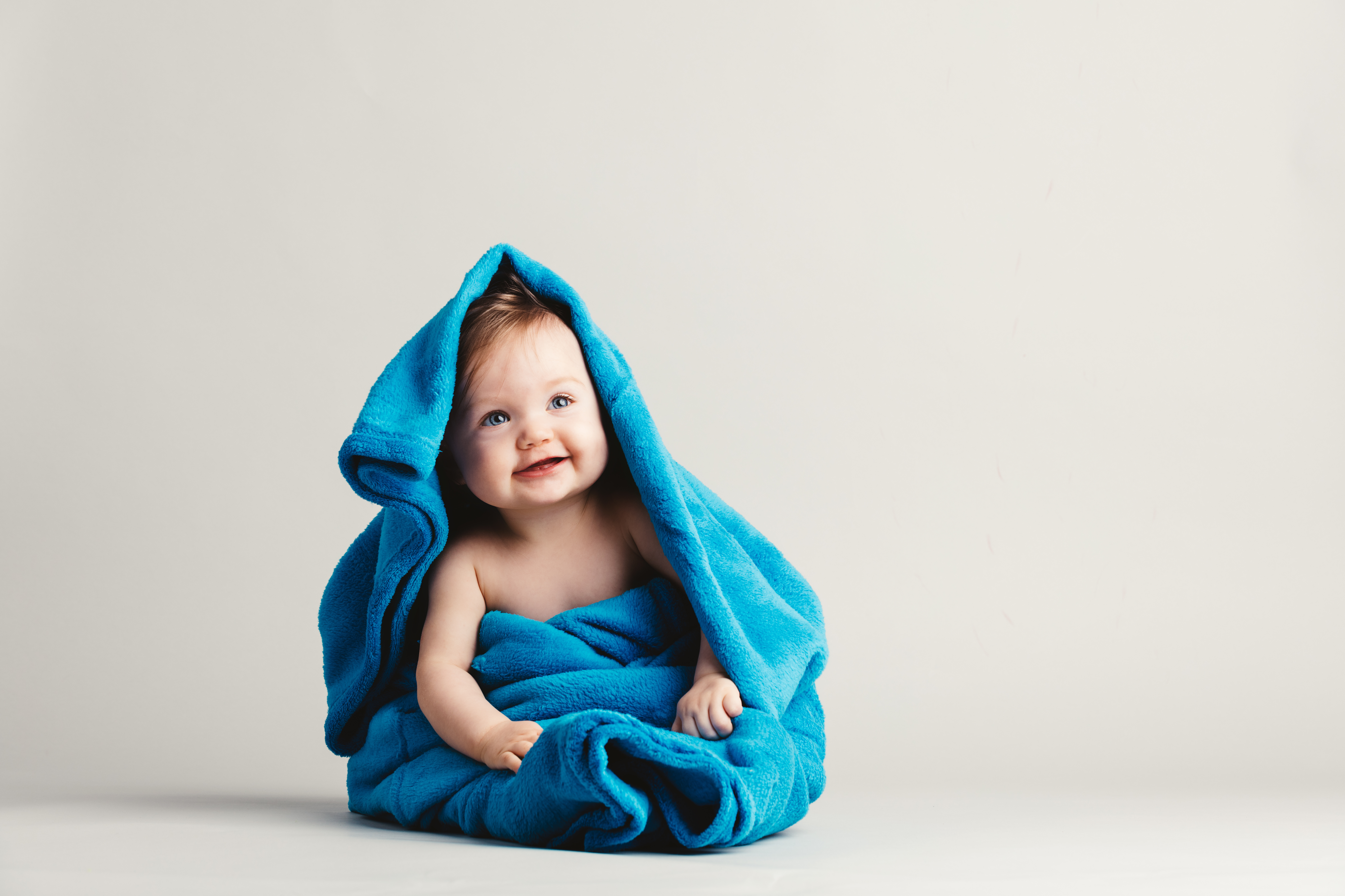 White backdrop with a blue towl wrapped around a smiling baby that has blue eyes.