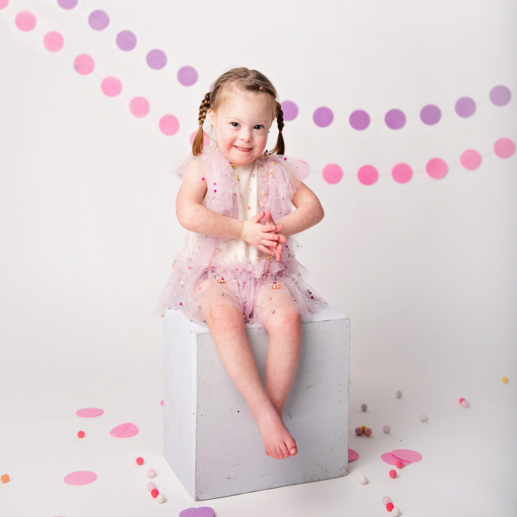 Young girl with Downs Syndrome sitting barfoot inon a white box.
