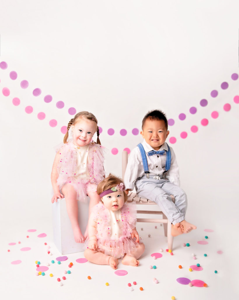 Three children with Downs Syndrome smiling in white studio wih colorful decorations.