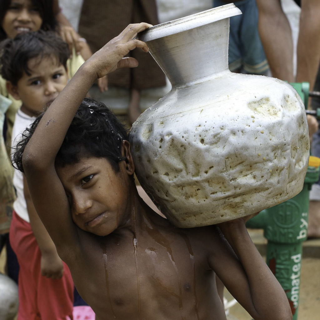 A shirtless young boy carries a larger silver container of water on his shoulder.