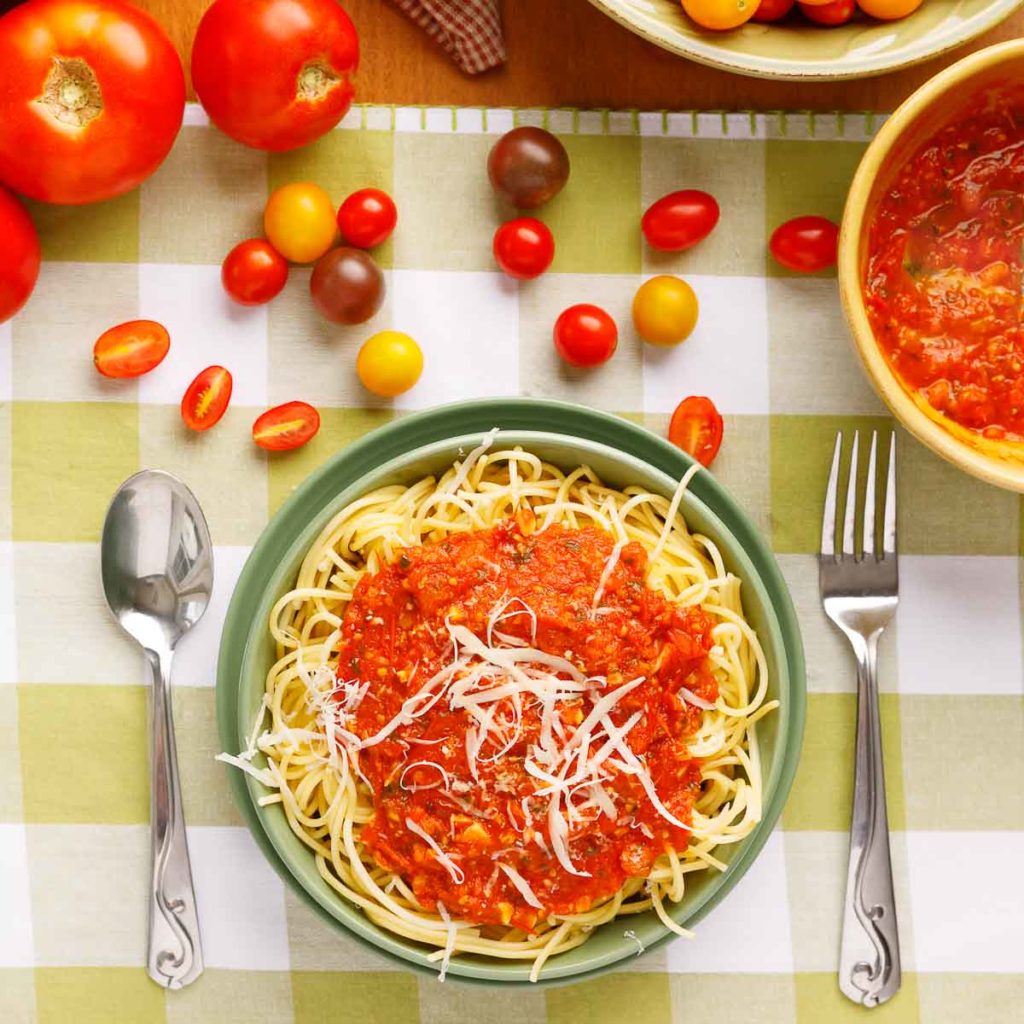 Top down view bow of spaghetti, spoon and fork, tomatoes, green and white checked place setting, bowl of spagetti sauce