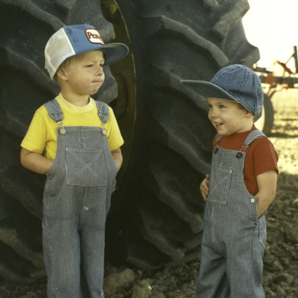 Two welker farms brothers standing by tractor tires when they were children, dressed in striped blue overalls and hats.