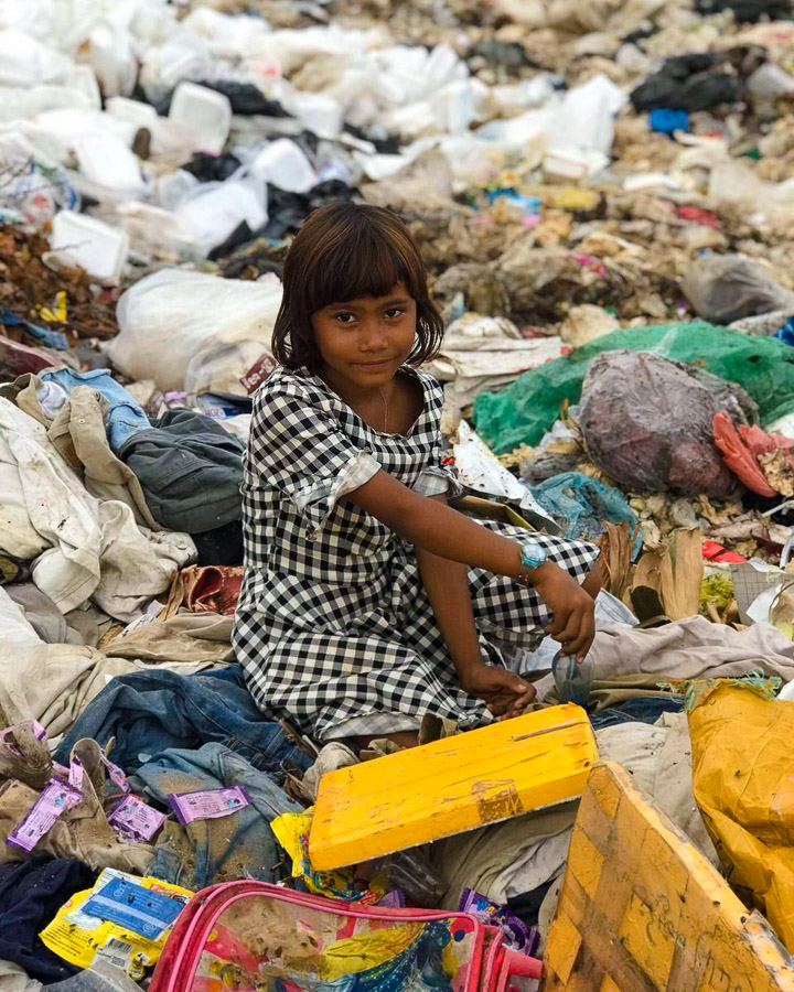 A young girl in a checkered dress pauses as she is sifting through a garbage pile.