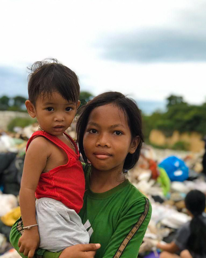 A young Cambodian girl stands in front of a garbage dump holding a young child.