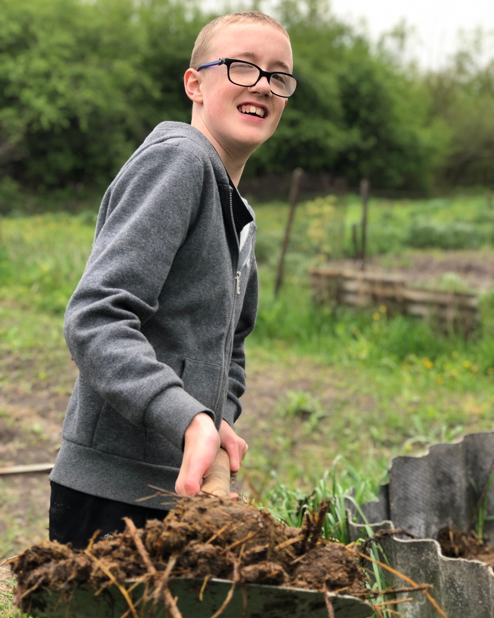 A young teenage boy in glasses and a grey sweatshirt squints as he turns and shovels compost for the family garden.