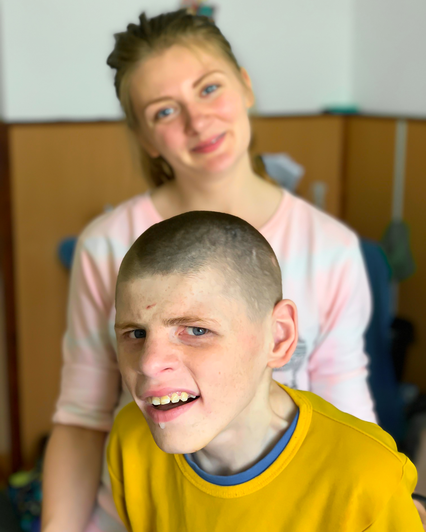 A teenage girl stands smiling behind a teenage boy with special needs in a brightly light room.