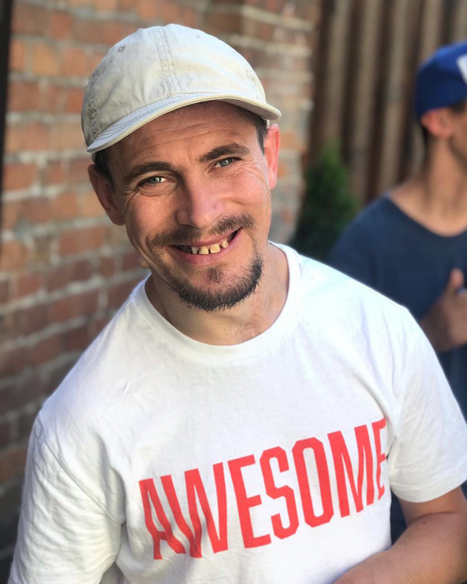 A special needs young man in his early 20's smiles wearing a white shirt with "awesome" written on it.