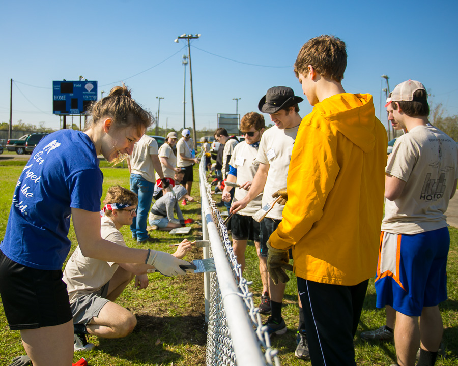 Eight Days of Hope volunteers paint both side of a chainlink fence near an athletic facility.