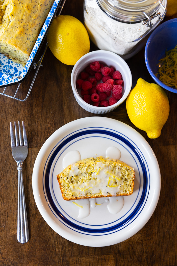 A slice of poppy seed bread drizzeled with frosting on a wooden table next to raspberries, lemons and made from scratch poppy seed mix.