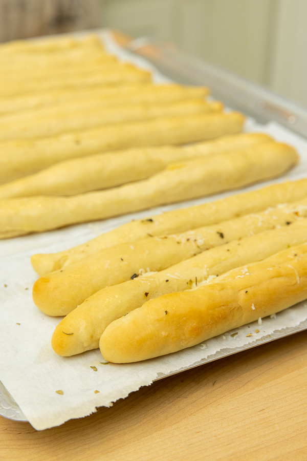 Pre-made breadsticks cooling on a flat pan, brushed with butter and drizzled with spices and grated cheese.