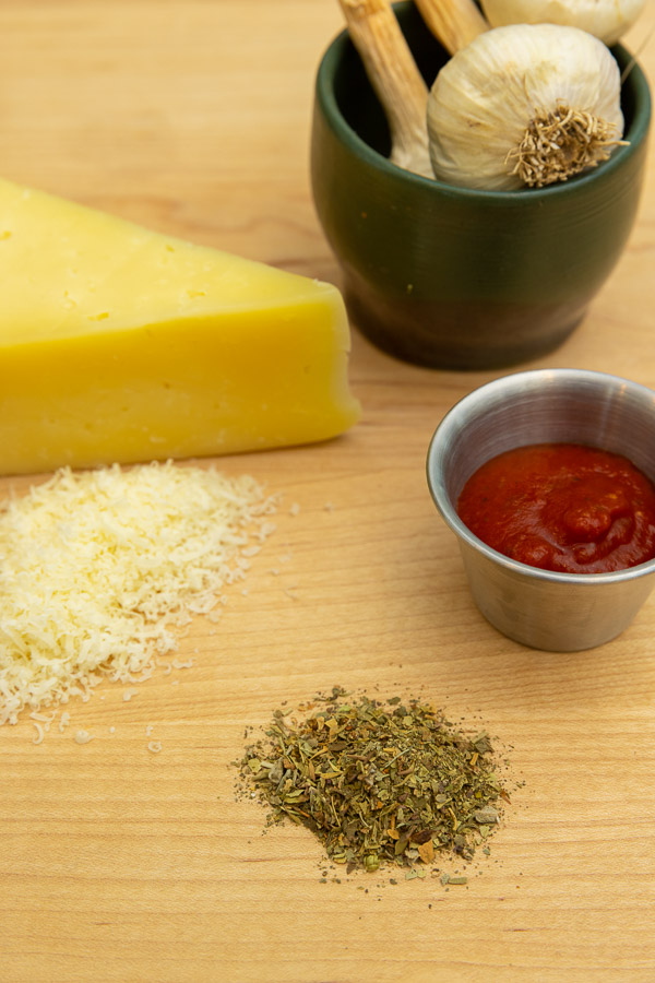 Cheese, spices, tomatoe sauce and a small bowl holding garlic cloves on a wooden countertop.