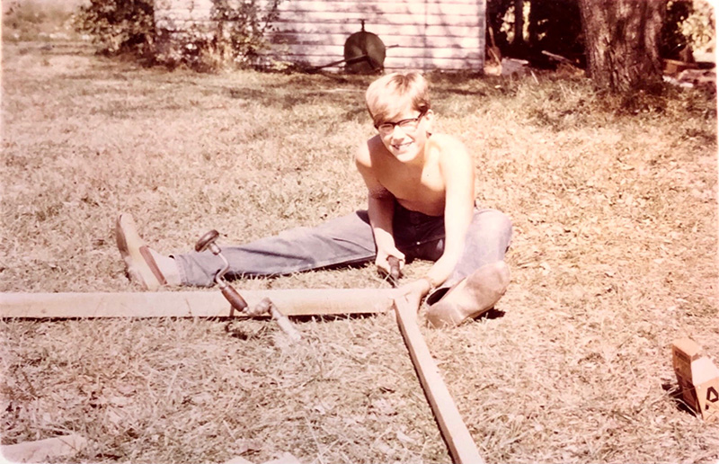 Joel Salatin as a boy, sitting on the ground building a chicken coop with hand tools.