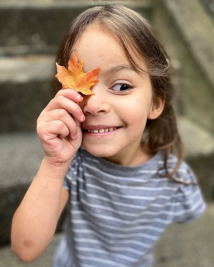 Zorey Gagnon smiles as she holds a orange and yellow leaf over her right eye.