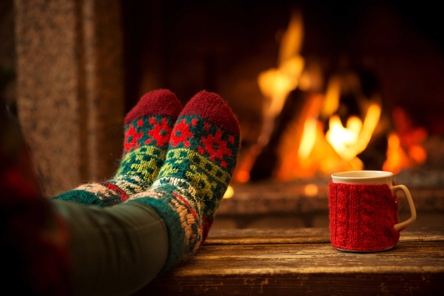 Someone relaxing in front of a fire with thier feet propped up on a wooden table, drinking coffee or hot chocolate.